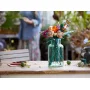 Bouquet de fleurs LEGO coloré dans un vase vert transparent posé sur une table en bois, avec une personne floue en arrière-plan Bouquet de fleurs LEGO coloré dans un vase vert transparent posé sur une table en bois, avec une personne floue en arrière-plan